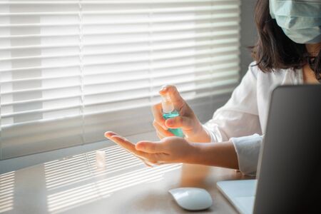 Closeup woman wear face mask using alcohol spray to clean hands on desk with laptop computer in office. Coronavirus diseases prevention concept.の写真素材
