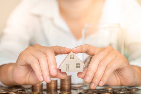 Closeup woman holding house figure with two hands with coins stack and glass jar on table. Business and savings money concept.の写真素材