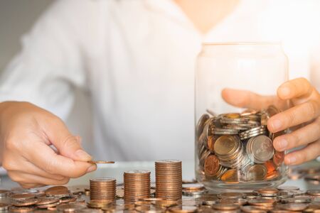 Closeup woman hand putting coin on stack with glass jar on table. Business and savings money concept.の写真素材