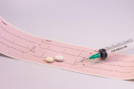 Electrocardiogram strip with a syringe and its needle and a pair of white pills. Free space to...