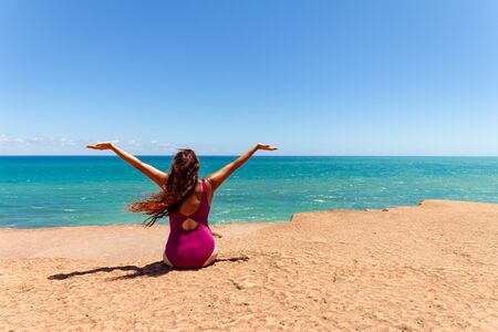 Happy woman facing the sea. Concept of happiness and joy. Woman with reddish hair and full color mesh board sitting on a cliff.の写真素材