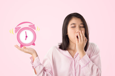 young asian woman wear pink pajamas yawn and holding alarm clock feeling sleepyの写真素材
