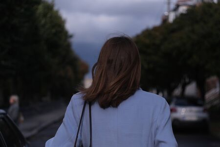 Young brunette woman crossing the traffic road in the evening and walking in the city centre alone,...