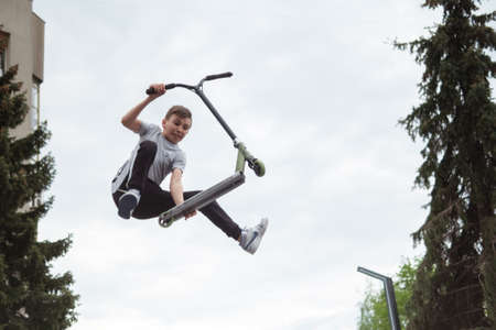 Zhytomyr, Ukraine - May 15, 2021: Teenager did unsuccessful trick on sportive push scooter on a city sports ground. He jumping over an obstacles. Extreme sports is popular among youth. Back rear viewのeditorial素材