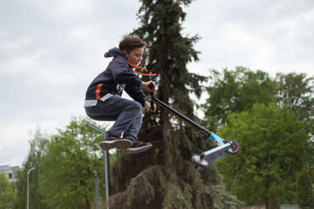 Zhytomyr, Ukraine - May 15, 2021: Teenager did unsuccessful trick on sportive push scooter on a city sports ground. He jumping over an obstacles. Extreme sports is popular among youth. Back rear viewのeditorial素材
