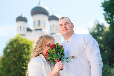 Zhytomyr, Ukraine - August 20, 2021: Couple in white shirt enjoying the summer. Attractive girlfriend sniffing bouquet of roses in the street at church backgroundのeditorial素材