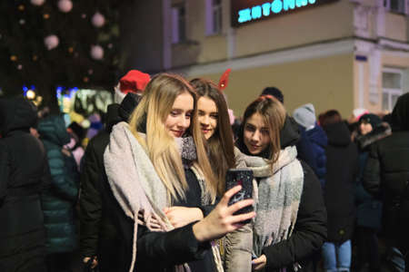 Zhytomyr, Ukraine - December 19, 2021: People gathered at night to celebrate the new year. Girls making selfie against christmas treeのeditorial素材