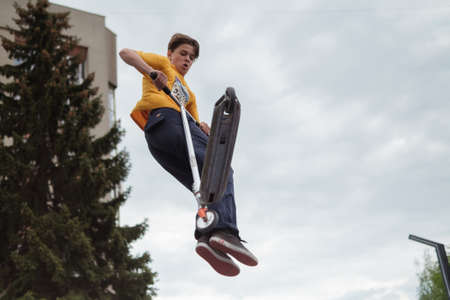 Zhytomyr, Ukraine - May 15, 2021: Teenager did unsuccessful trick on sportive push scooter on a city sports ground. He jumping over an obstacles. Extreme sports is popular among youth. Back rear viewのeditorial素材