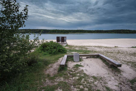 Simple wooden bench by a lake with beautiful view on a calm water surface and lake shore. Relaxing atmosphere. Nobody. Dramatic cloudy skyのeditorial素材