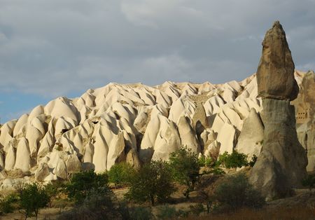Horizontal composition of grey sky, a chine of fantastic white tuff formations and dark-green trees in the foreground. As for the central object we see it on the right: it's a typical for Cappadocia mushroom shaped stone formation.の写真素材