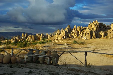 A composition of three different coloured parts. From the top: 1. blue sky with dark-grey clouds, 2. the chine of tuff formations alight with sunset, 3. a sort of fence and a row of national clay pitchers in the shade の写真素材