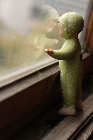 Very old (about 50-s) baby-doll in pale-green overalls is half-face standing by the muddy window (the weak reflection of its face visible in the glass) の写真素材