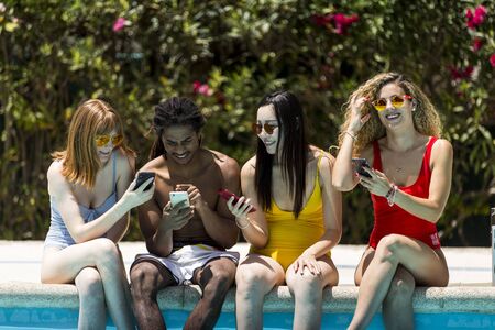 Happy boys and girls in the pool consulting their smartphones. Multiracial group of friends sitting at the edge of the pool share images with your mobile phone.の写真素材