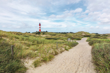 Tall red and white striped lighthouse building standing on small hill. Sandy coastal landscape with green vegetation and footpaths. Summer day at seaside.の写真素材