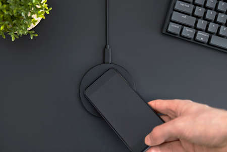High angle view of dark gray office desk. Male hand putting smartphone on wireless charger. Modern...
