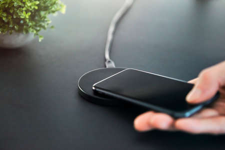 Man placing smartphone on induction wireless charger in office. Gray desk surface background. Modern technology, wireless device and transfer of energy concept.の写真素材