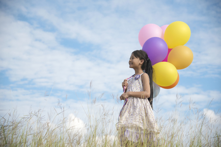 Happy girl holding colorful of balloons on a green meadow with cloudy and blue skyの写真素材