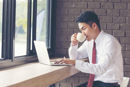 Asian Handsome businessman working outdoor in the coffee shop with laptop and holding cup of coffee on hand. Work outside office using technology with wifi, wireless connection. Technology Concept.の写真素材