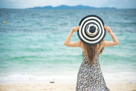 Summer time tan woman vacation on the beach. Cheerful woman wear summer dress and straw hats sitting on the beach look at sea. Time to relax in summer lifestyle outdoor shot on tropical island beach.の写真素材