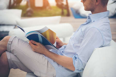 Caucasian young man reading magazine book on sofa reader holding a book open paper page and reading while sitting outdoor on sofa out of library and reading book. Lifestyle for man.の写真素材
