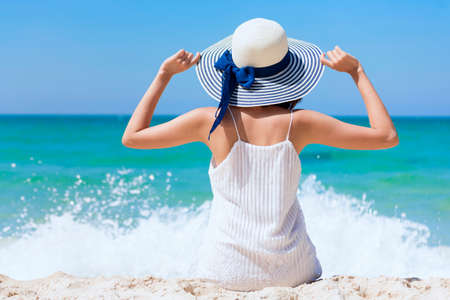 Summer time woman vacation on the beach. Cheerful woman wear summer dress and straw hats sitting on the beach look at sea. Time to relax in summer lifestyle outdoor shot on tropical island beach.の写真素材