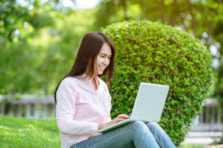 Asian woman sitting green park using laptop computer. Woman working on laptop happy entrepreneur business using notebook with hands typing on keyboard home office during coronavirus quarantine periodの写真素材