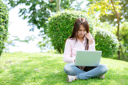 Asian woman sitting green park using laptop computer. Woman working on laptop happy entrepreneur business using notebook with hands typing on keyboard home office during coronavirus quarantine periodの写真素材