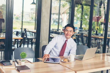 Businessman using tablet in cafe via wireless internet technology working online. Asian handsome young man holding smart tablet using gadget with handheld and laptop on table. Freelance Lifestyleの写真素材
