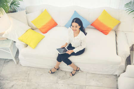 side view of Young beautiful Asian woman look at the camera sitting on white sofa using tablet smartphone explore website. reading article and online content.の写真素材