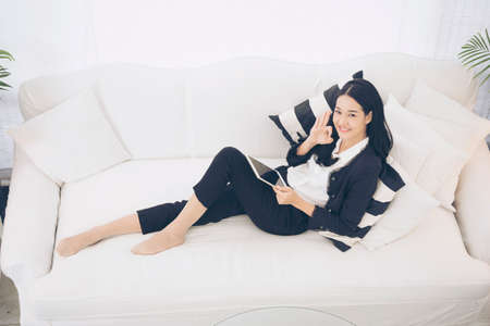 side view of Young beautiful Asian woman look at the camera sitting on white sofa using tablet smartphone explore website. reading article and online content.の写真素材