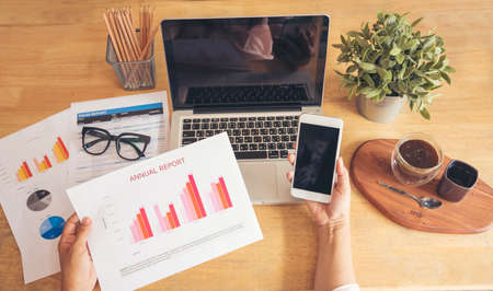 Woman working at home office desk using laptop business financial document chart and graph on wooden table with coffee cup. Woman freelance reading business graphs sale report on business office deskの写真素材