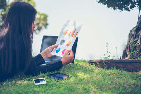 Asian entrepreneur business woman reading business graph analyze chart using laptop on green garden at home. Freelance working home office social distance new normal lifestyle in open cafe green parkの写真素材
