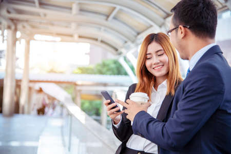 Businessman Businesswoman drinking coffee in town using smartphone outside office modern city. Hands holding take away coffee cup and smart phone talking together Business partner with cup of coffeeの写真素材