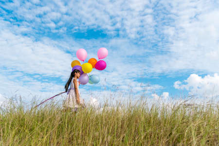 Cheerful cute girl holding balloons running on green meadow white cloud and blue sky with happiness. Hands holding vibrant air balloons play on birthday party happy times summer on sunlight outdoorの写真素材