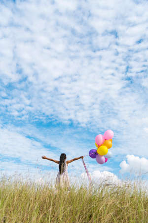 Cheerful cute girl holding balloons running on green meadow white cloud and blue sky with happiness. Hands holding vibrant air balloons play on birthday party happy times summer on sunlight outdoorの写真素材