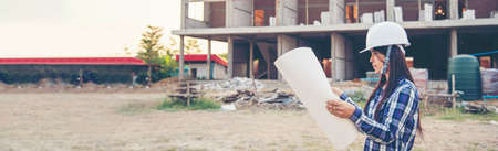 Banner Woman construction engineer hold blueprint wear plaid shirt safety white hard hat at construction site industry. Panorama Architecture Female civil engineer look at blueprint with copy spaceの写真素材
