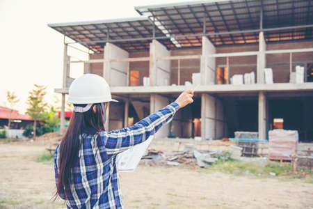 Woman construction engineer hold blueprint wear plaid shirt safety white hard hat at construction site industry labor worker. Architecture Female engineer civil worker look at blueprint real estateの写真素材