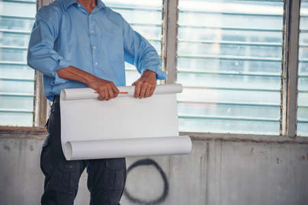 Construction engineer man hold blueprint wear blue shirt safety white hard hat at construction site industry labor worker. Architecture man engineer civil worker look at blueprint real estate conceptの写真素材