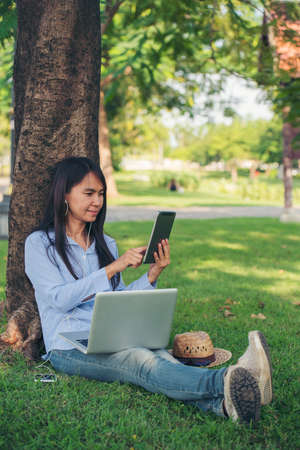 Asian woman sitting green garden park using phone listen music from smart phone with laptop...