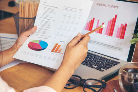 Business woman using smartphone, laptop on office desk. Female freelance reading financial graph charts Planning analyzing marketing data. Hands of young woman working in office firm business info.の写真素材