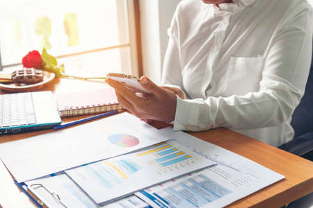 Business woman using smartphone, laptop on office desk. Female freelance reading financial graph charts Planning analyzing marketing data. Hands of young woman working in office firm business info.の写真素材