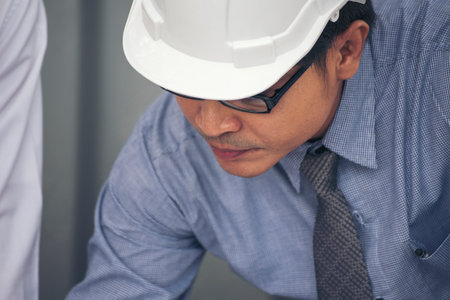 Civil construction engineer working with laptop at desk office with white yellow safety hard hat at office on construction site. Asian young man architecture project manager sitting at office on siteの写真素材