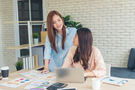 Two asian Female freelance reading financial graph charts Planning analyzing marketing data. Asian people team working office firm. Two businesswomen team meeting using laptop at company office desk.の写真素材