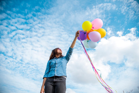 Woman holding balloons running on green meadow white cloud and blue sky with happiness Cheerful and relax. Hands holding vibrant air balloons play on birthday party happy times summer sunlight outdoorの写真素材