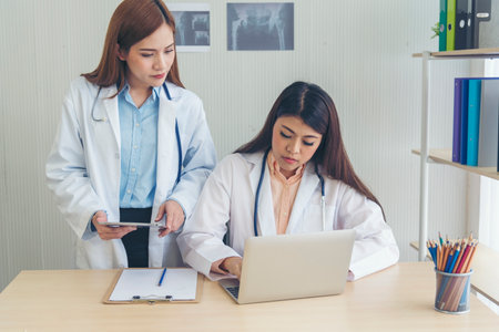 Two asian women doctors discuss meeting doctor's office medical clinic using laptop consulting patient disease. Asian medical lab young women talking together discussing healthcare assistance teamworkの写真素材