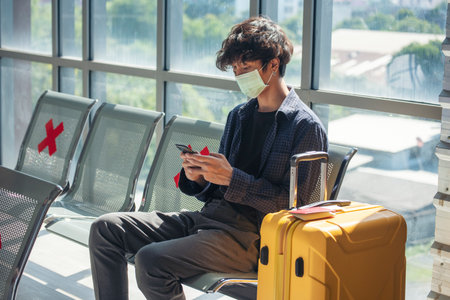 Asian young man traveller new normal wearing face mask sitting Social distancing holding smartphone in Airplane lounge. New normal Male travel pandemic passenger by plane. Coronavirus tourism tripの写真素材