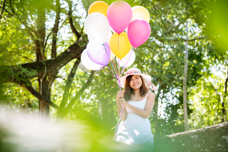 Cheerful beauty woman holding balloons relax sitting under big tree in green park with happiness. Woman Hands holding vibrant air balloons play on birthday party happy time summer on sunshine outdoorの写真素材