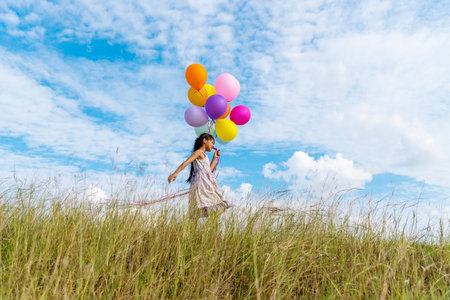 Cheerful cute girl holding balloons running on green meadow white cloud and blue sky with happiness. Hands holding vibrant air balloons play on birthday party happy times summer on sunlight outdoorの写真素材