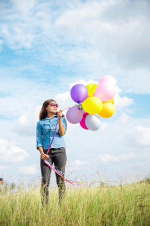 Woman holding balloons running on green meadow white cloud and blue sky with happiness Cheerful and relax. Hands holding vibrant air balloons play on birthday party happy times summer sunlight outdoorの写真素材