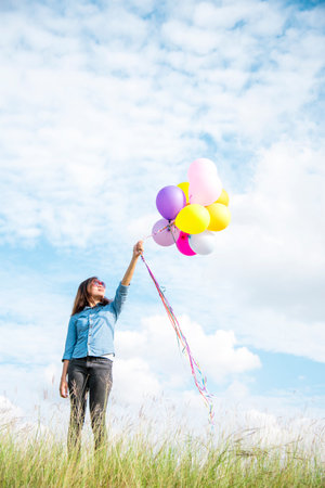 Woman holding balloons running on green meadow white cloud and blue sky with happiness Cheerful and relax. Hands holding vibrant air balloons play on birthday party happy times summer sunlight outdoorの写真素材